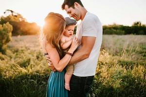 A young couple holding a baby in what looks to be a field, with the sunshine in the background, everyone is very  happy